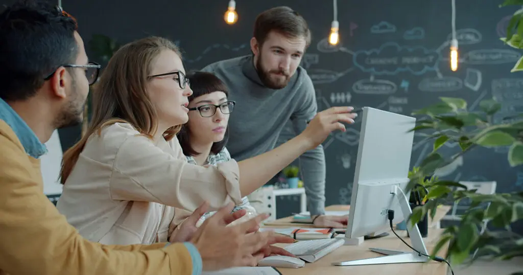 Young people colleagues are talking and laughing indoors in open space office enjoying work together sitting at desks and pointing at laptop screen.